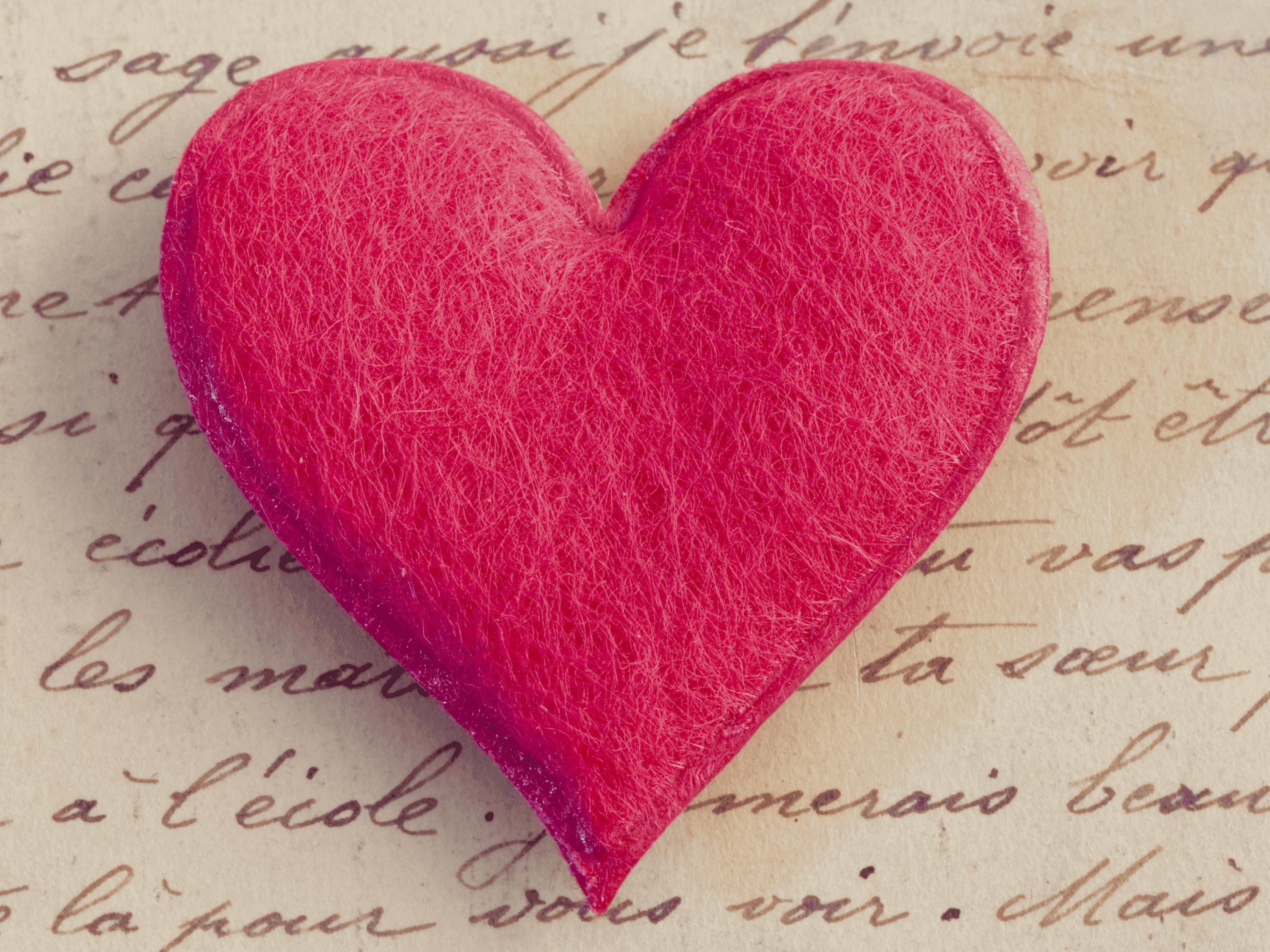 Ein leuchtend rotes, herzförmiges Objekt mit auffälliger Textur befindet sich in der Mitte eines Hintergrunds aus altem Papier mit eleganten, handgeschriebenen Buchstaben, die eine romantische oder nostalgische Stimmung vermitteln.
